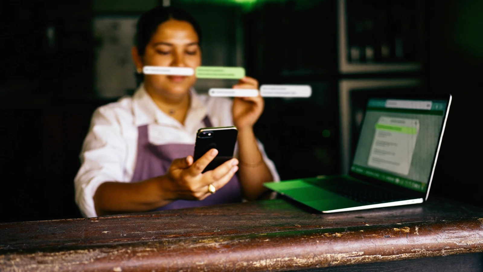 Dona de loja respondendo mensagens de clientes pelo celular com o notebook aberto no balcão